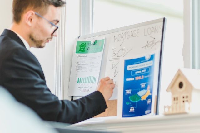 Person in a suit and glasses writing on a white board that reads