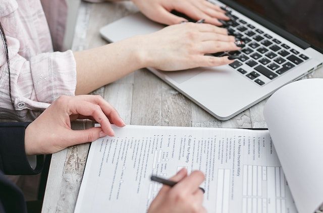 One pair of hands typing on a computer, another set of hands writing on a printed paper on a clipboard