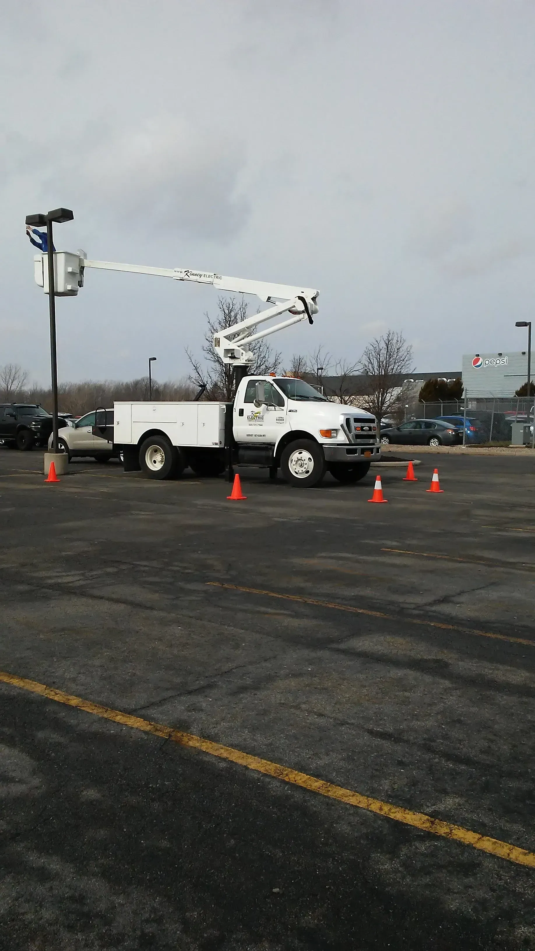 White utility truck with raised bucket arm in a parking lot, surrounded by orange cones under a gray sky