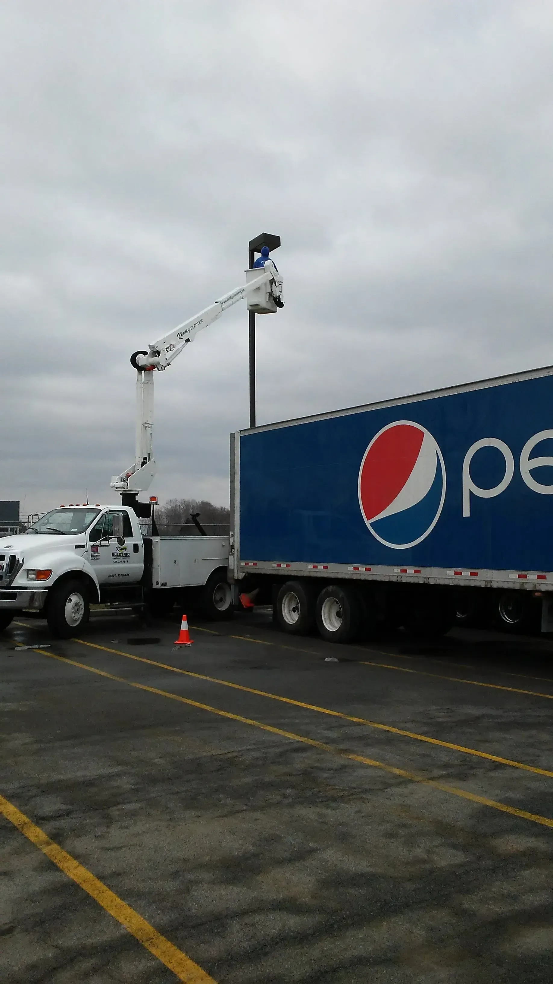 Utility truck and bucket lift beside a Pepsi trailer in a parking lot under a cloudy sky