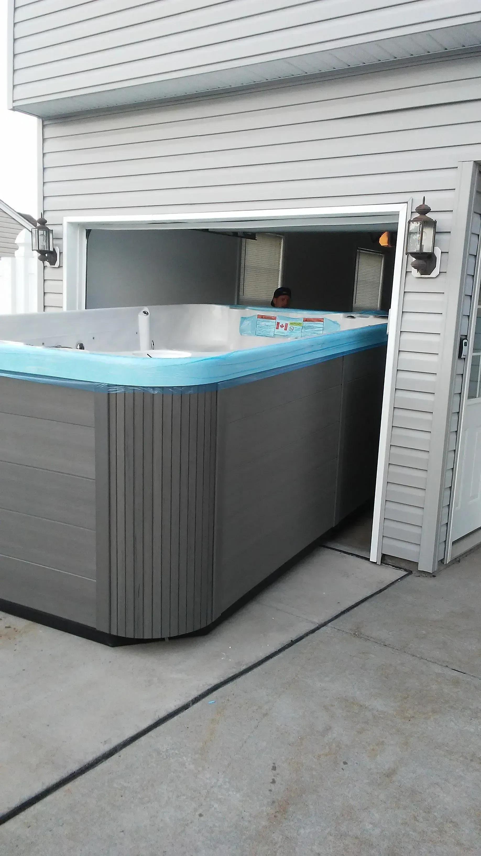 Outdoor hot tub in driveway beside a garage, with blue water and gray siding.