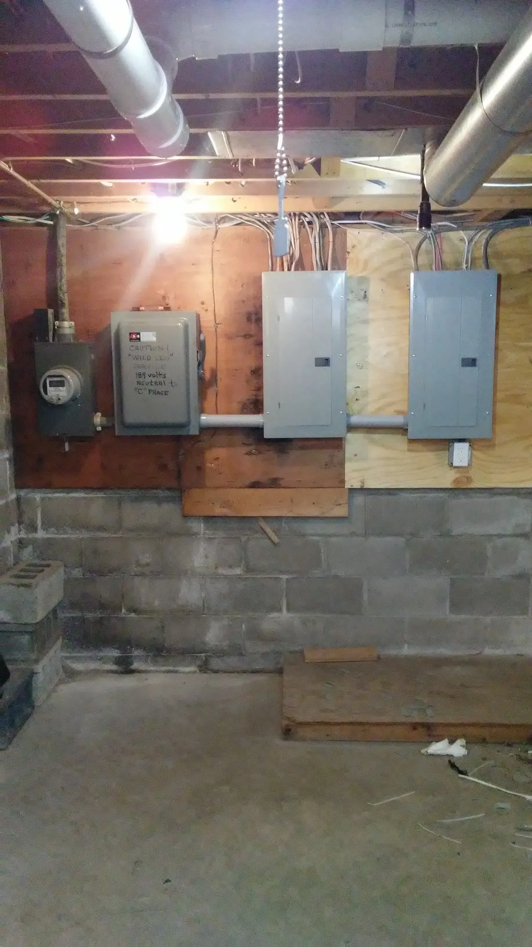 Basement utility wall with electrical panels, exposed pipes, and unfinished concrete floor