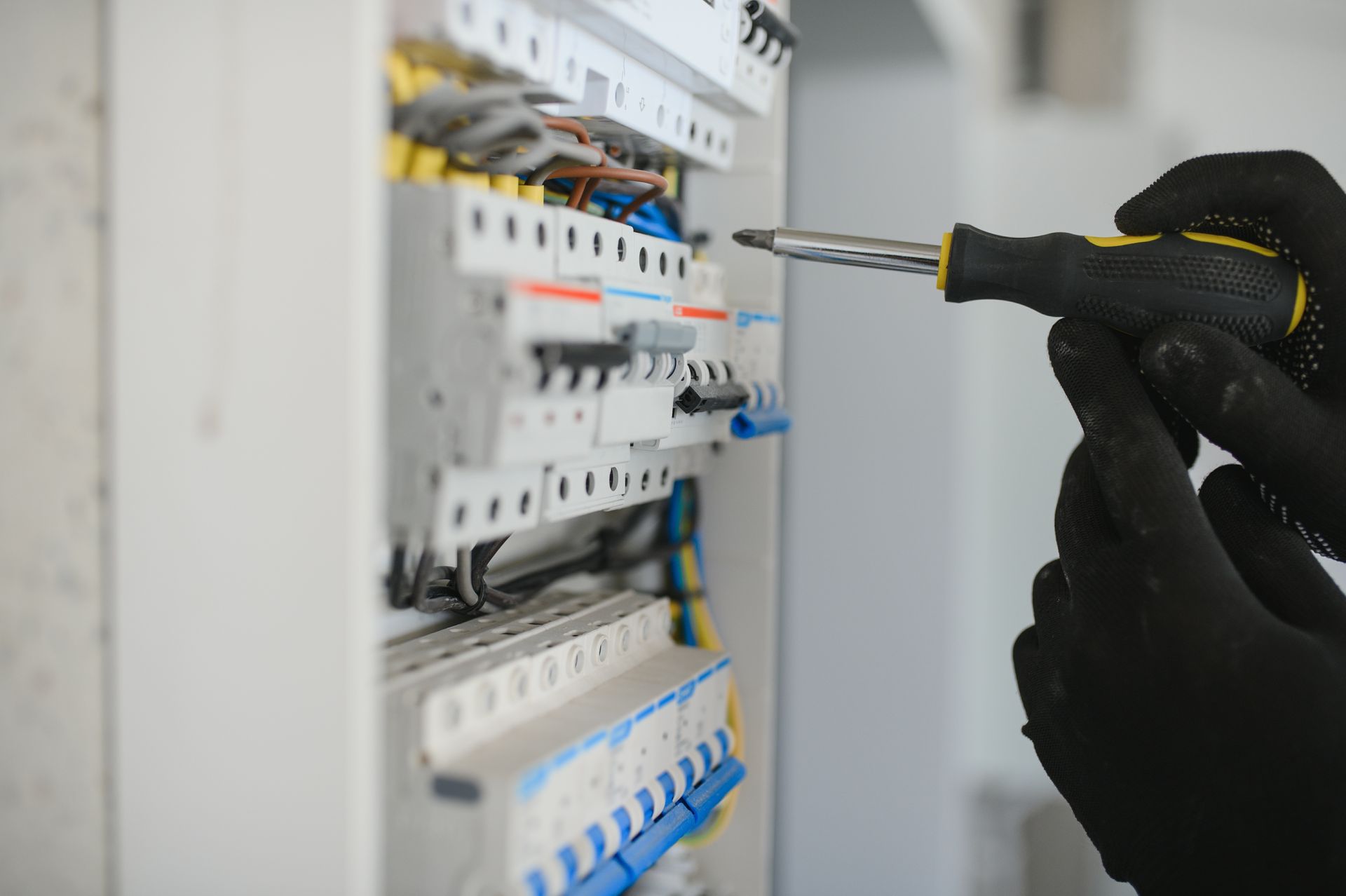Gloved hands adjusting wiring in an electrical breaker panel with a screwdriver.
