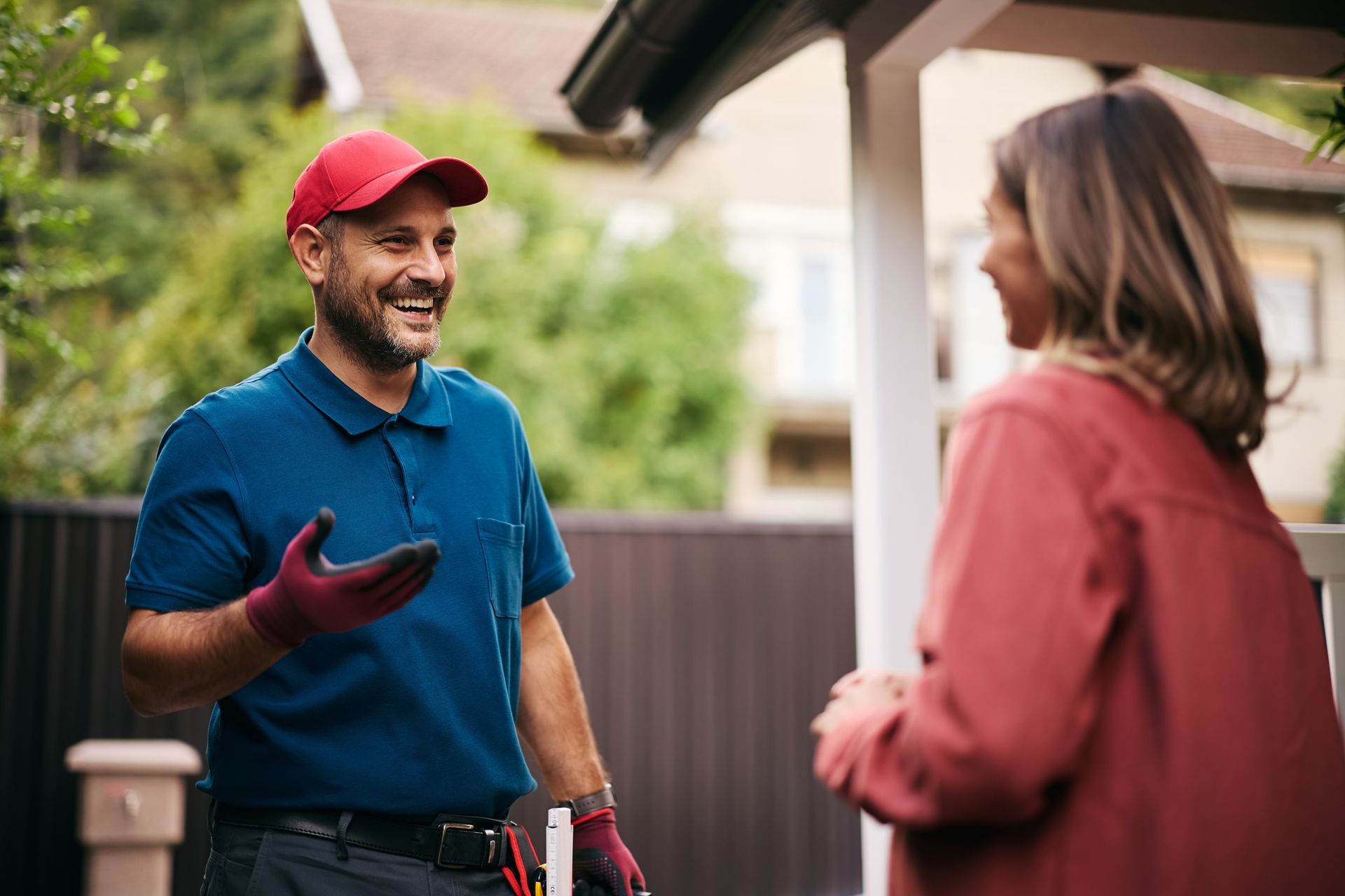 Man in red cap and blue polo talking and smiling with a woman on a porch.