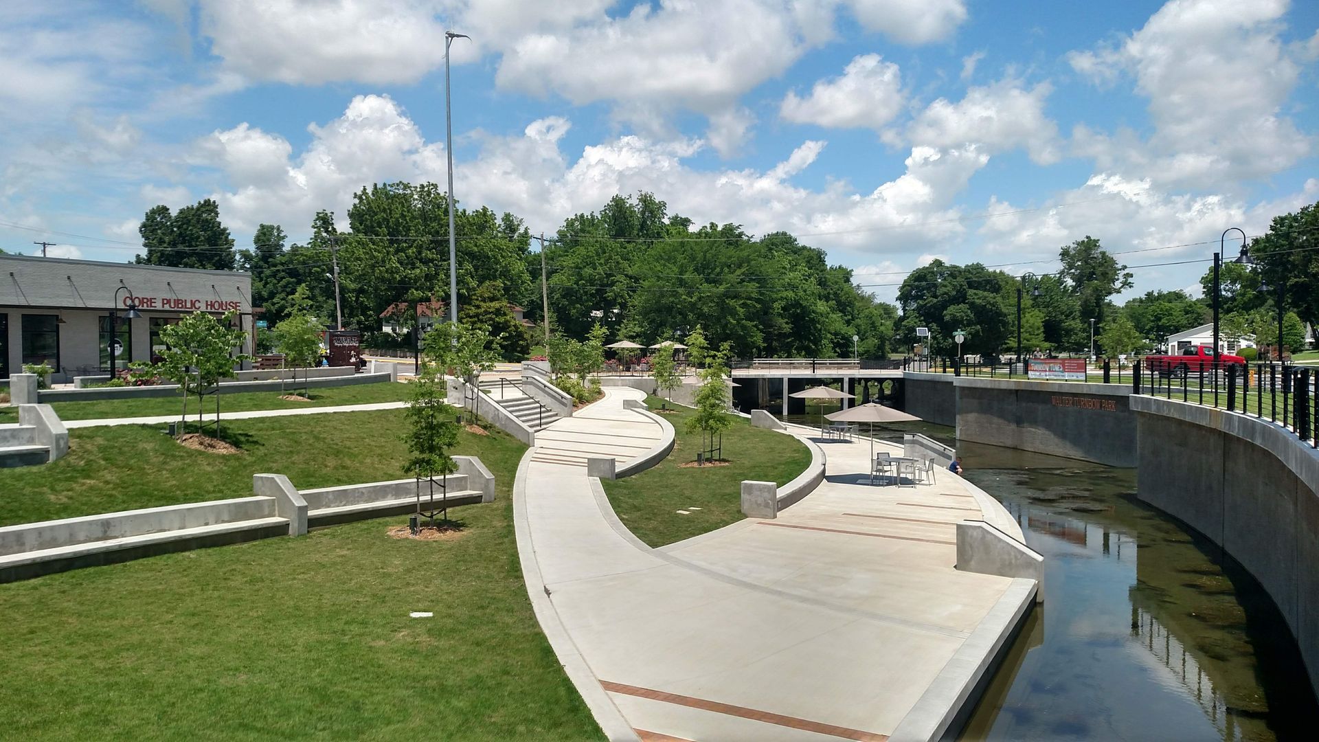 Curved riverside walkway and park with concrete benches, green lawn, trees, and cloudy blue sky