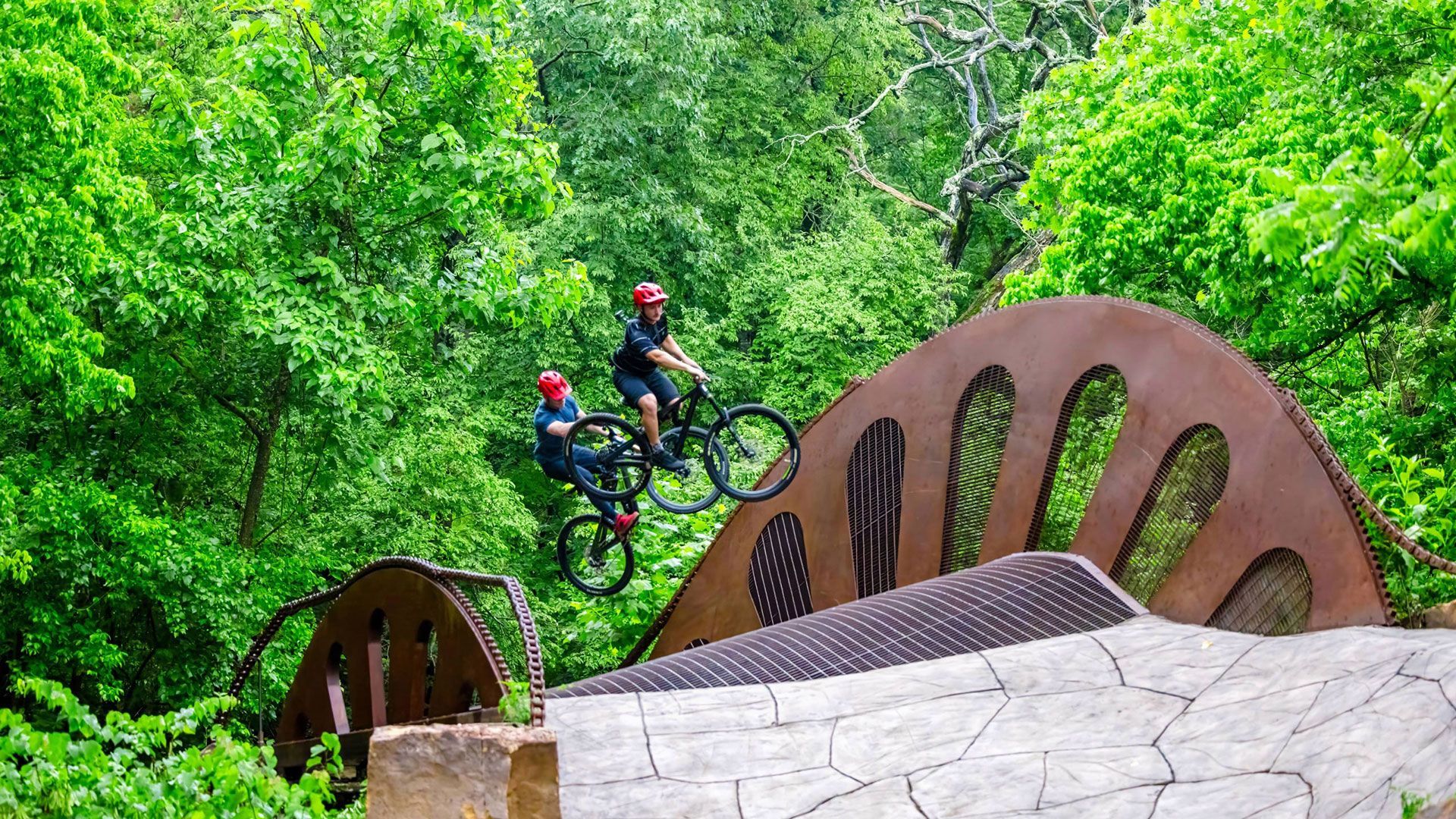 Two mountain bikers jump over a stone bridge in a lush green park