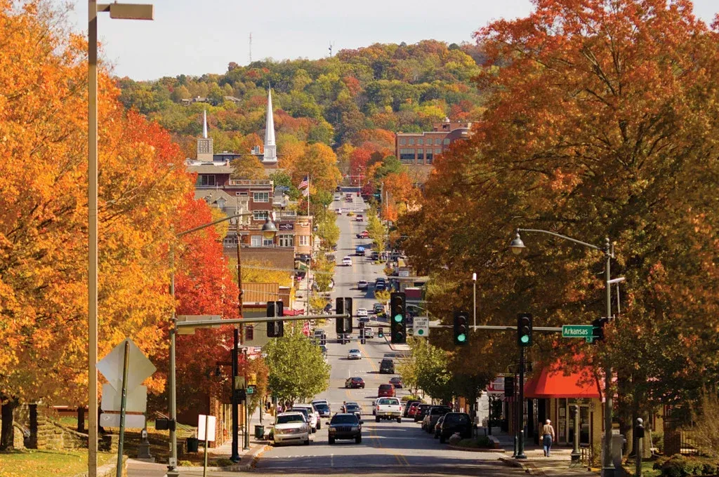 Tree-lined city street with autumn foliage, traffic lights, and hills in the background.