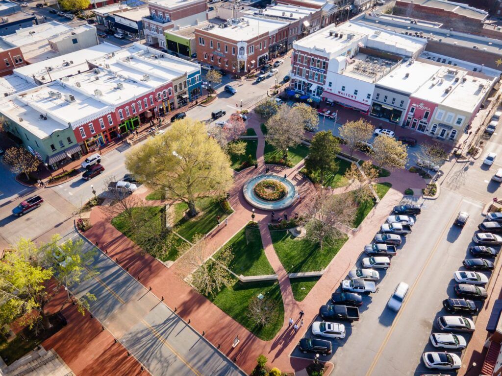 Aerial view of a town square with a fountain, pathways, trees, and surrounding buildings with parked cars.