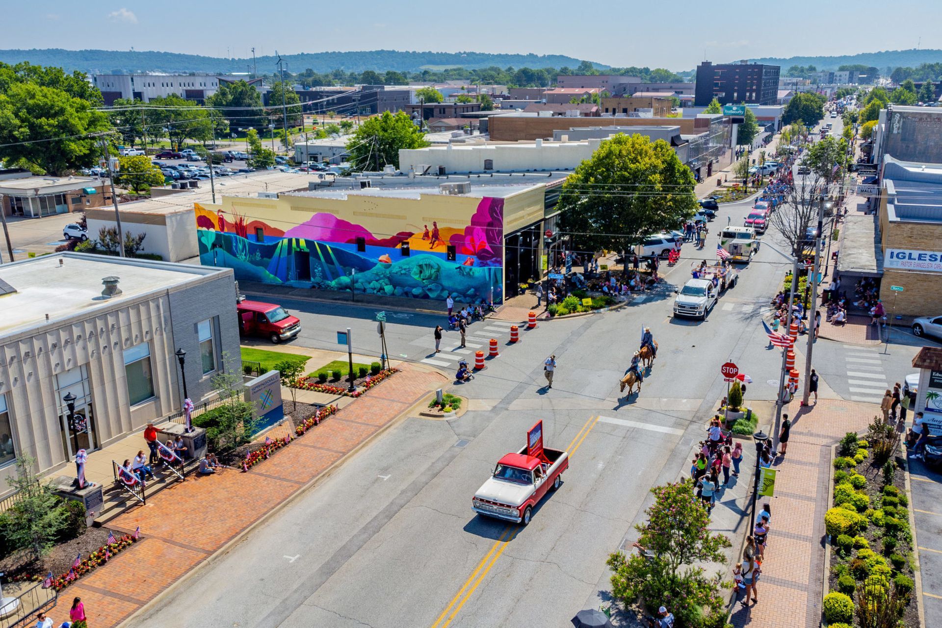 Aerial view of a lively street parade with colorful mural, marching participants, and parked cars on a sunny day