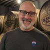 Smiling bald man with glasses in a brewery, standing before wooden beer barrels