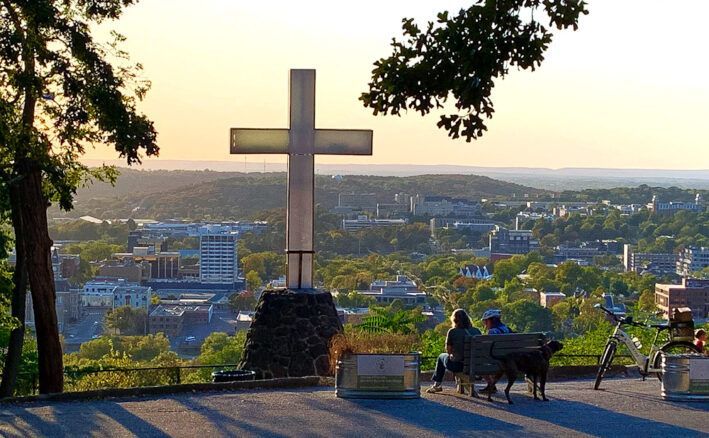 Large cross overlooking a city at sunset, with a bench and two people on a hilltop overlook
