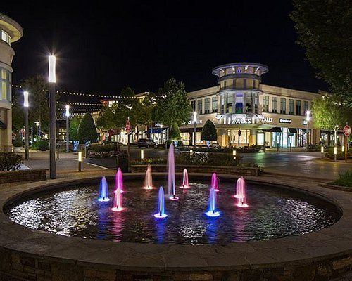 Nighttime plaza with colorful fountain jets and lit storefronts around a circular water feature