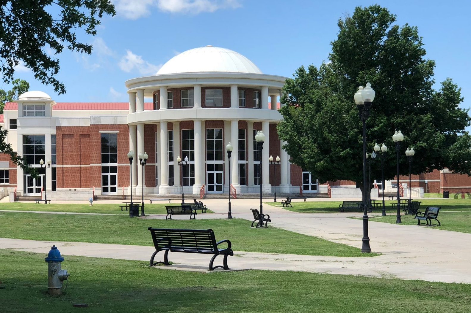 Brick campus building with white columns and dome beside a grassy plaza and benches under a blue sky