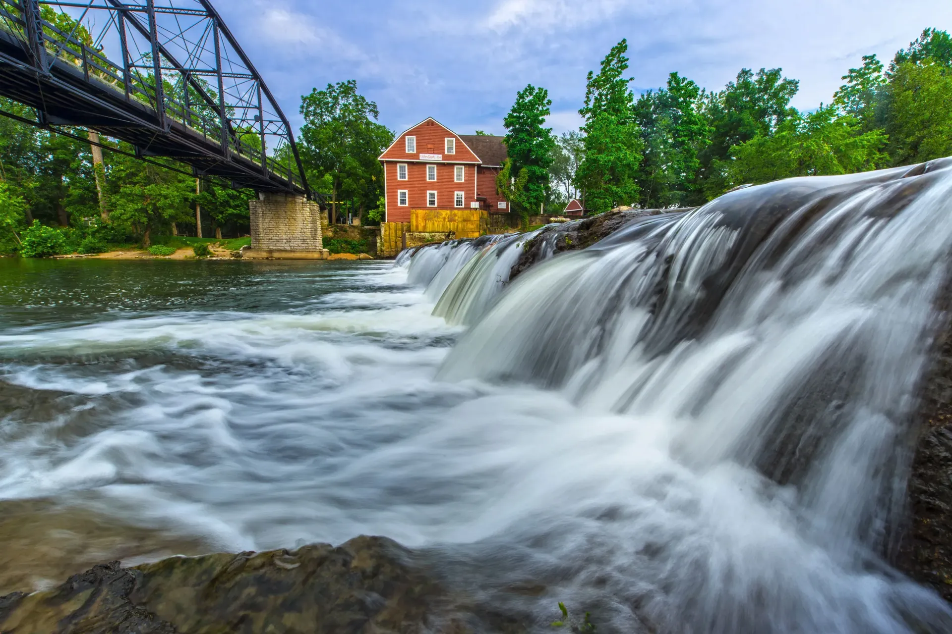 Waterfall beside a red mill and bridge in a lush green riverside landscape