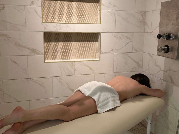 Person lying face down on a massage table in a tiled spa room, covered with a white towel.