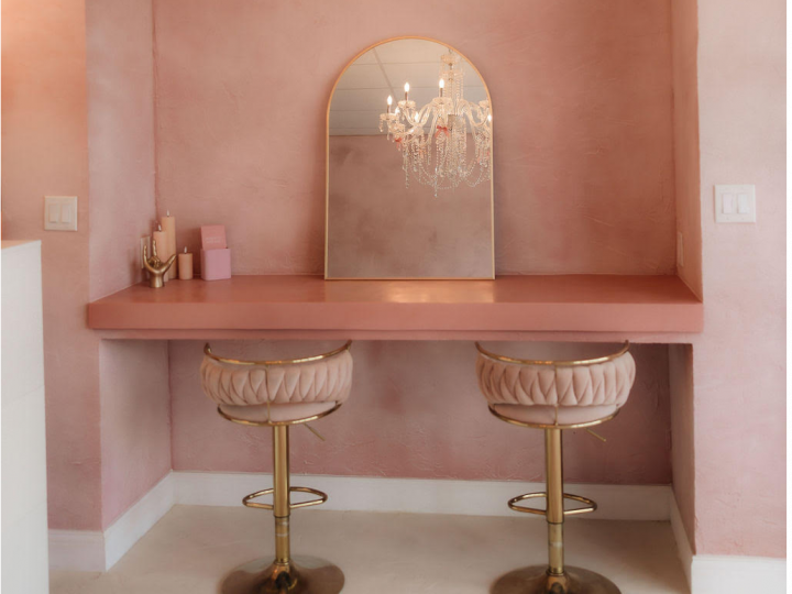 Pink vanity nook with floating desk, arched mirror, and two blush velvet stools