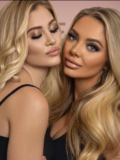 Two blonde women posing closely together with glamorous makeup against a pink background