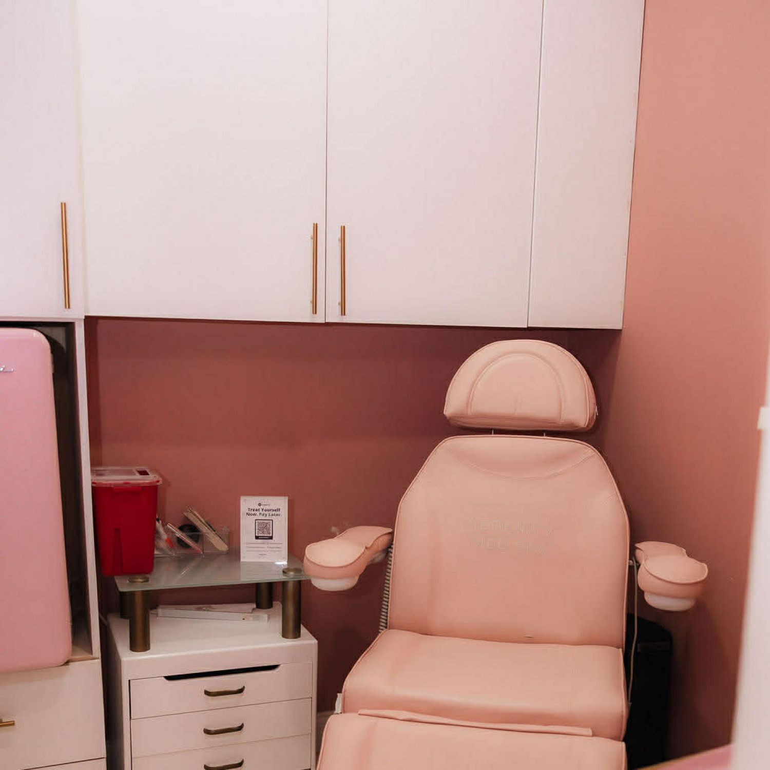 Pink salon chair beneath white cabinets in a small treatment room.