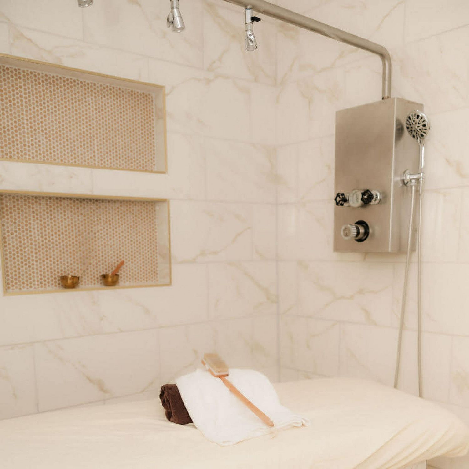 Person in a spa treatment room lying face down on a massage table under a towel, with a wall-mounted shower unit nearby