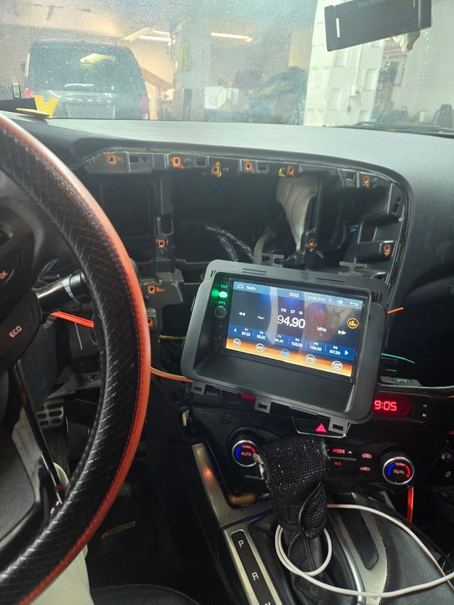 Car dashboard with a new radio display, wires exposed, partially disassembled. Steering wheel visible on left.
