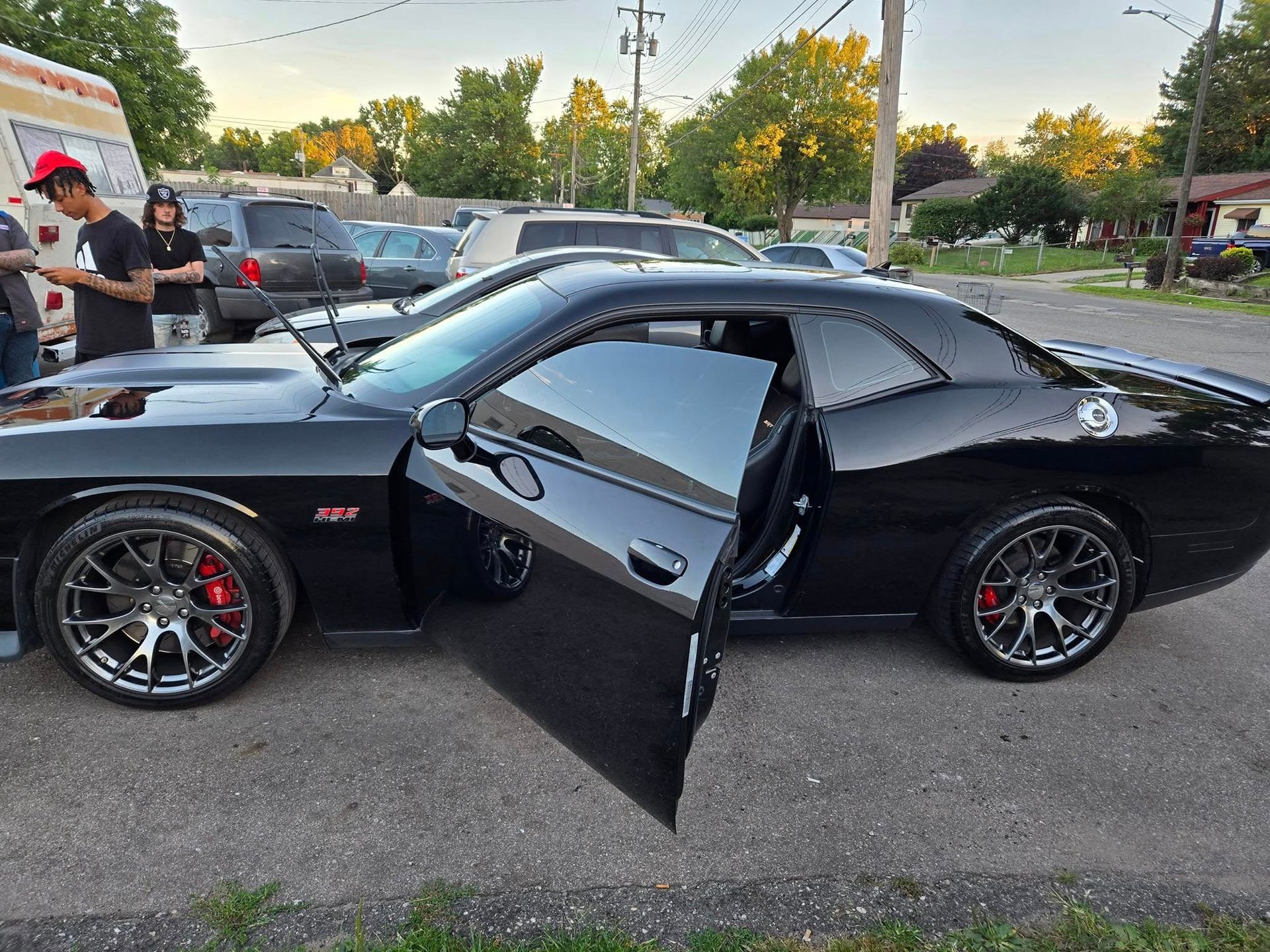 Black Dodge Challenger with open door parked on the street. People stand nearby.