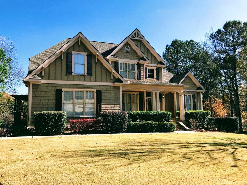 Large tan suburban house with a front porch, trimmed hedges, and a sunlit lawn under a blue sky