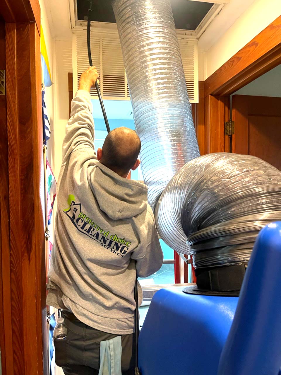 A man is hanging a air duct cleaning hose from the HVAC ceiling vent  in a room.