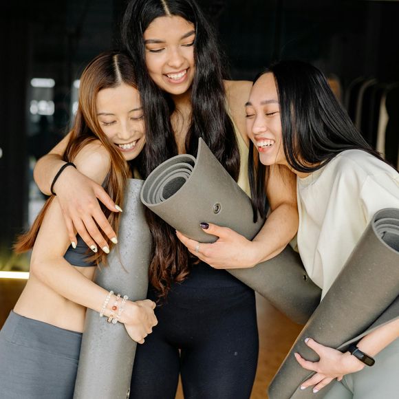 Three smiling people embrace, each holding a rolled yoga mat in a studio setting.