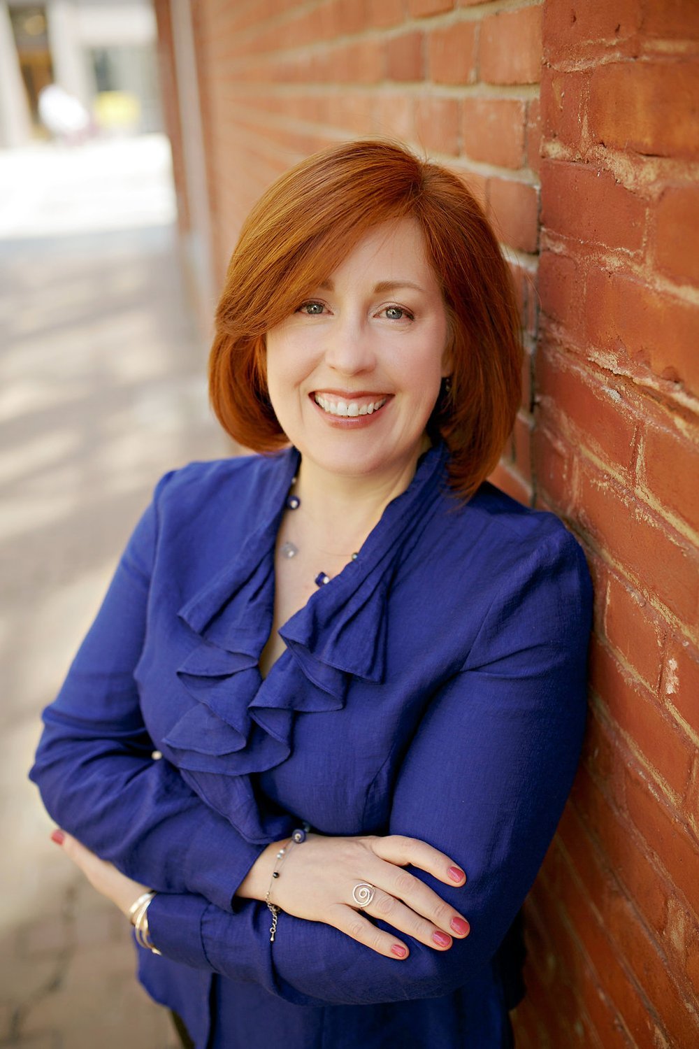 A woman is leaning against a brick wall with her arms crossed and smiling.