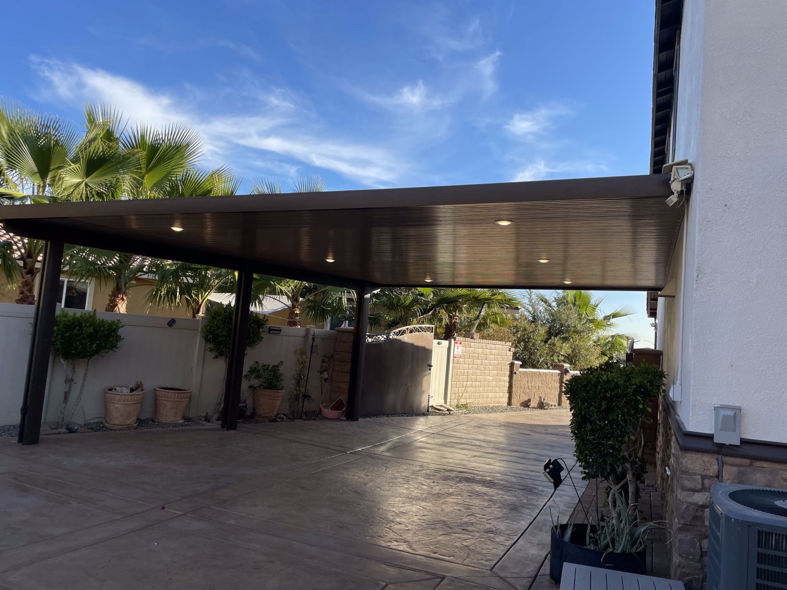 Carport attached to a house with a brown roof and posts. Concrete driveway, cloudy blue sky.