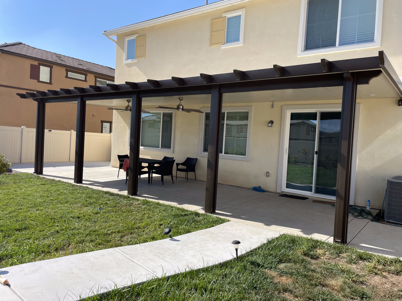 A backyard patio with a dark pergola, concrete path, and lawn.