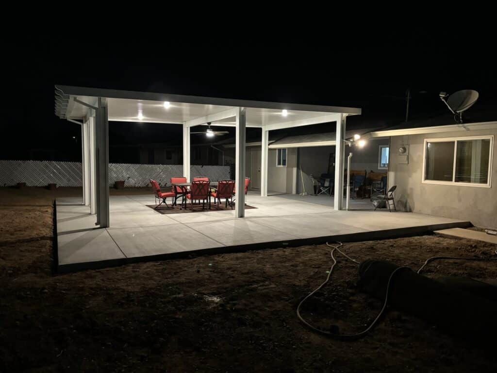 Backyard patio with red chairs and table under a lit white covered area. Dark night setting.