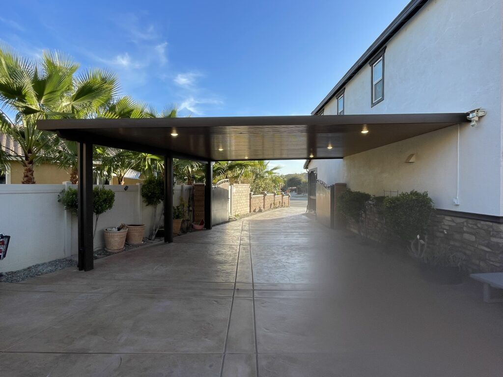 Brown carport attached to a two-story building. Concrete driveway. Palm trees and clear sky.