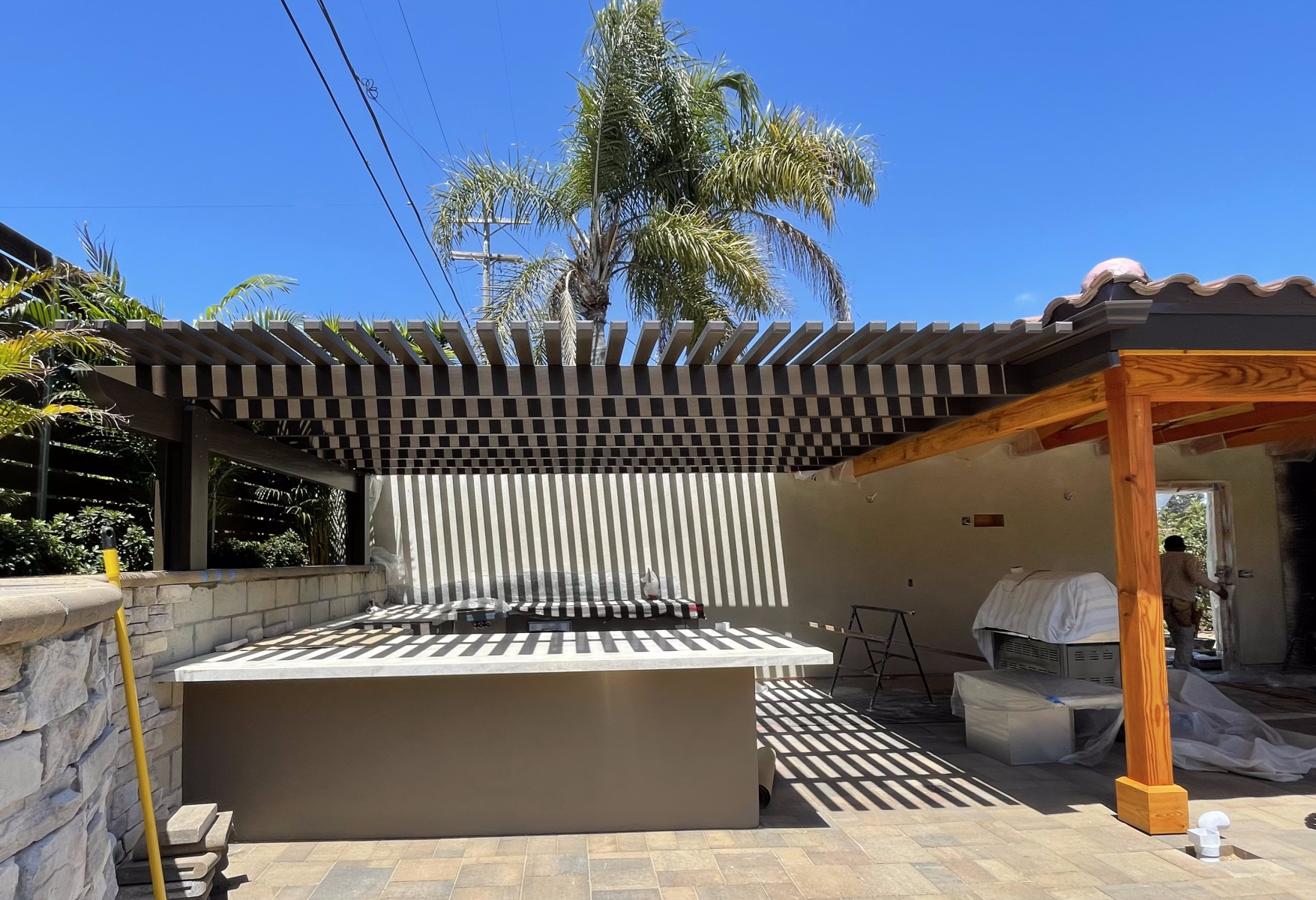 Outdoor patio with a dark slatted pergola casting shadows over a countertop and a palm tree in the background.