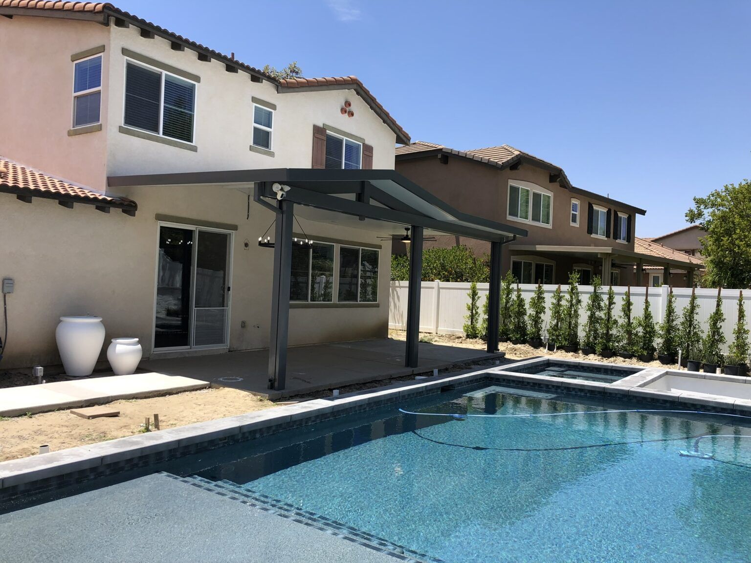 Backyard with a pool, patio cover, and two-story house. Blue water, white fence, and sunny day.