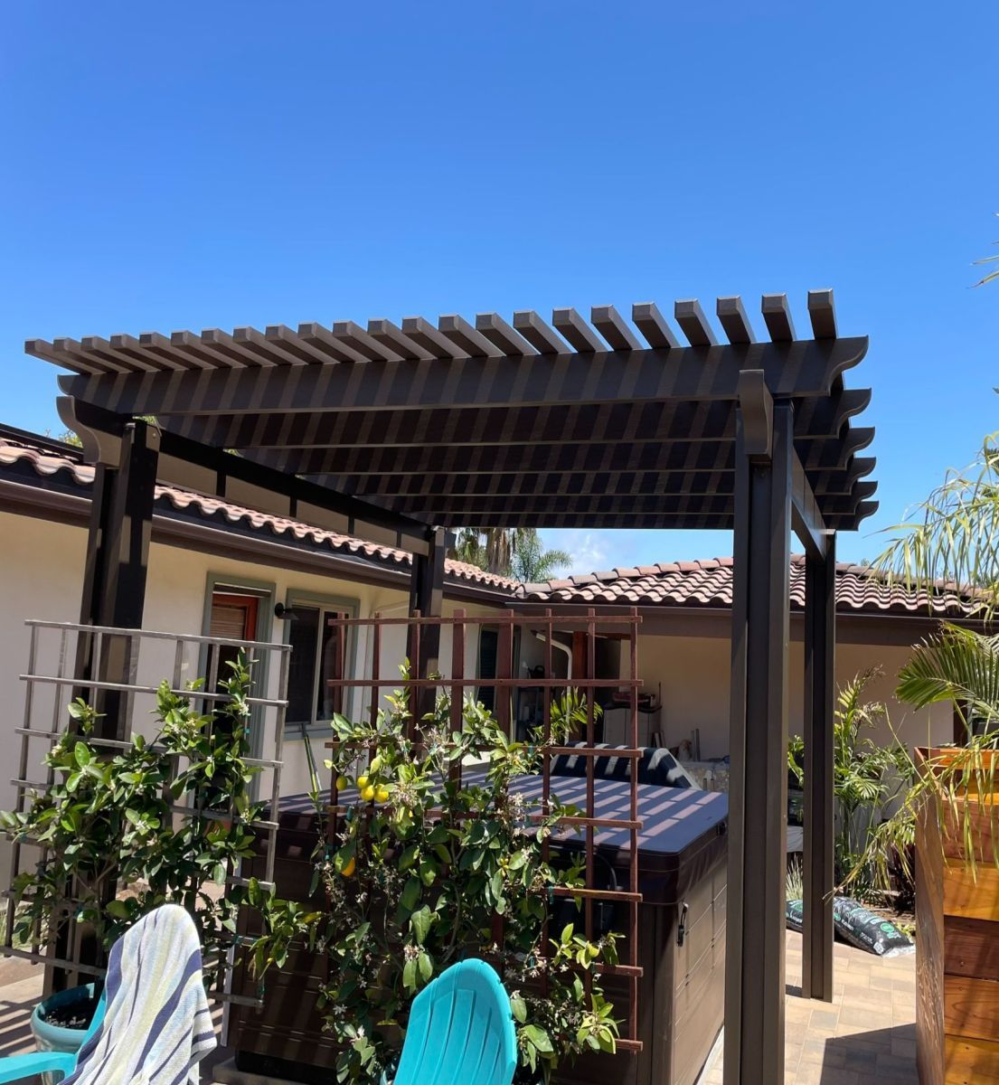 Brown pergola over a hot tub, with greenery and blue sky.