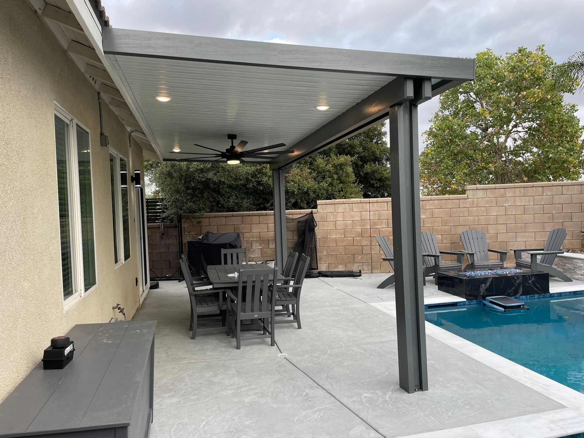 Patio with aluminum cover, dining table, pool, and a fire pit. Cloudy day.
