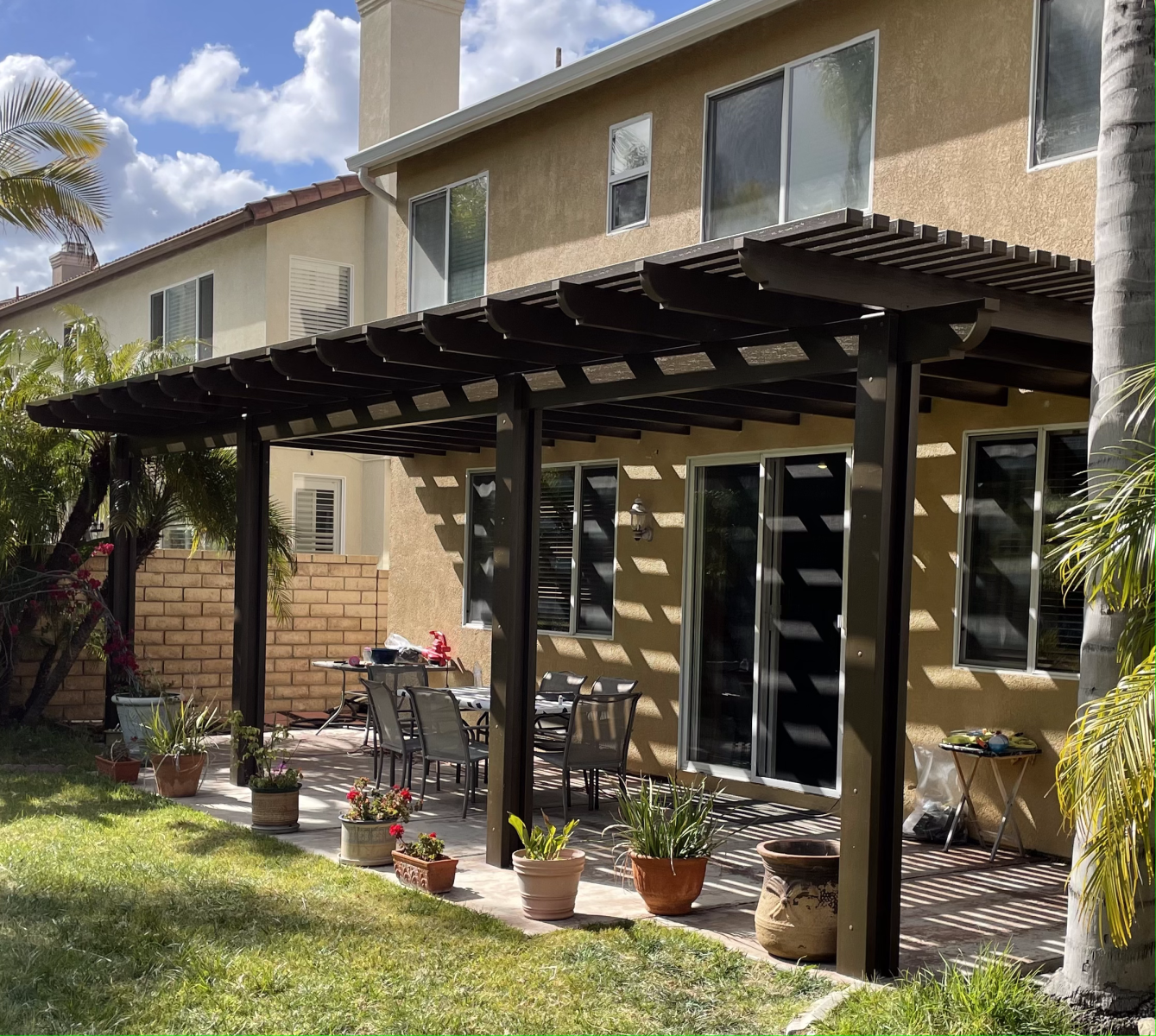 Backyard with pergola over a patio, plants, table and chairs. Beige house with multiple windows.