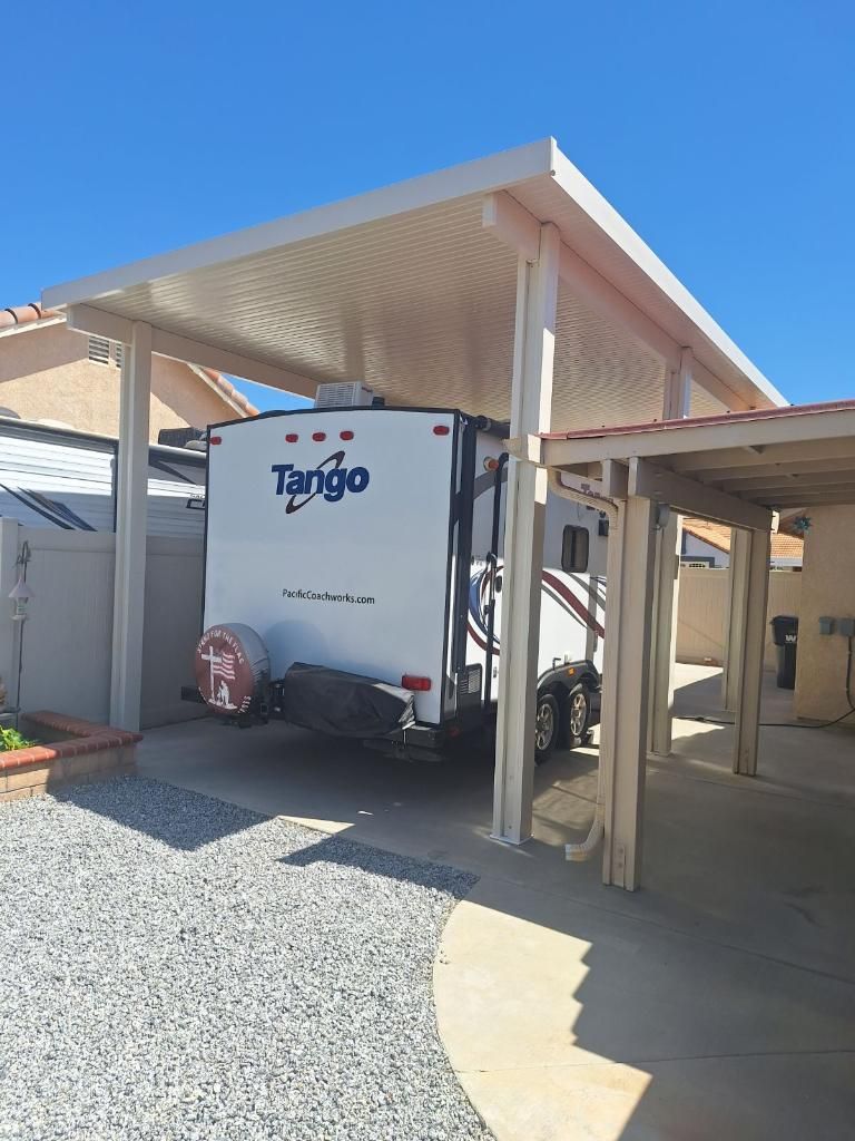 RV parked under a white metal carport in a gravel driveway. Another carport is adjacent. Blue sky.