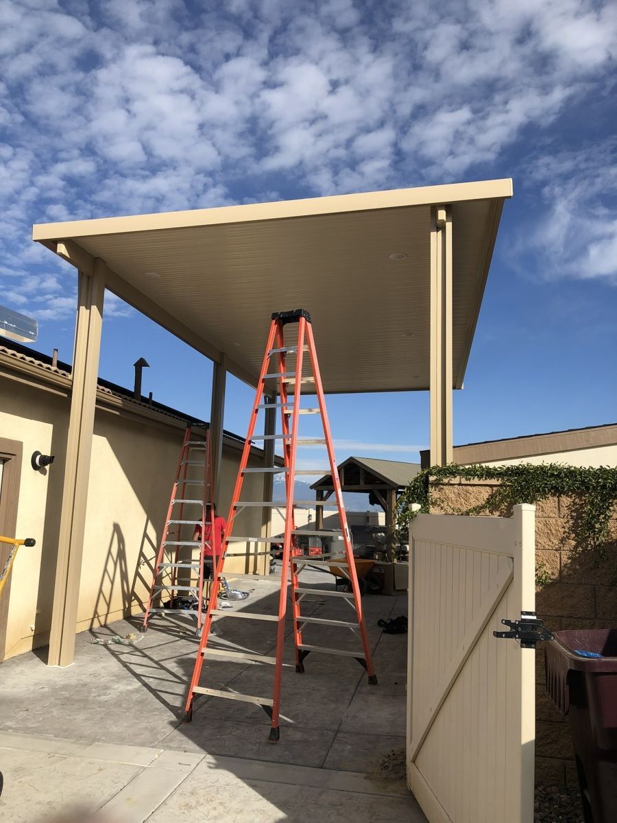 Patio cover with two ladders; a person near one. Beige and tan construction on a sunny day.