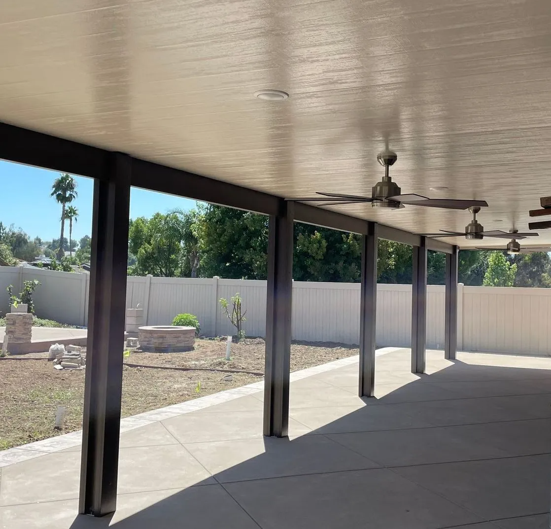 Covered patio with ceiling fans, dark supports, concrete floor, overlooking a yard with a white fence.