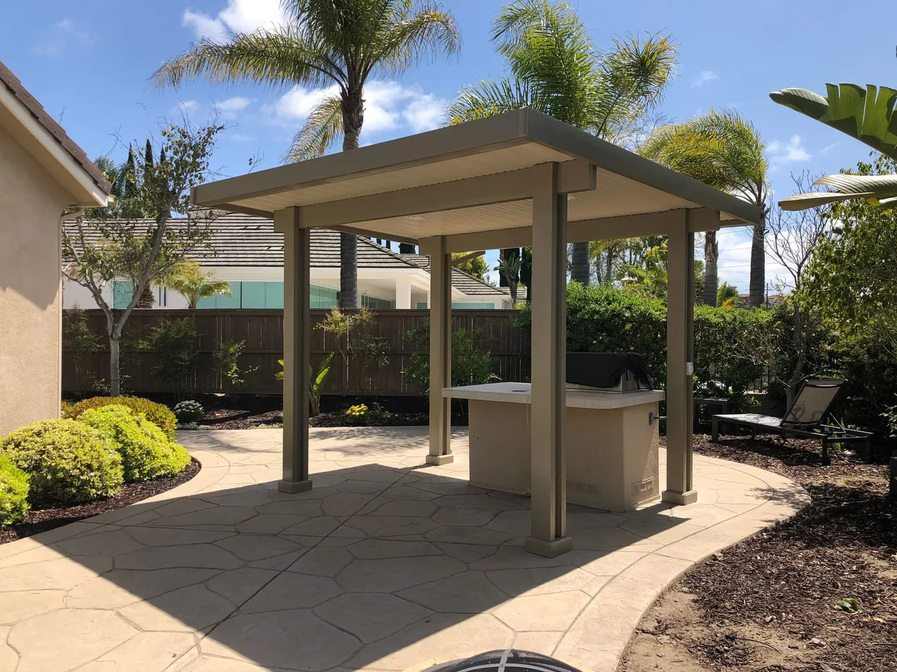 Beige pergola over a concrete patio with a built-in grill, surrounded by greenery and a fence.