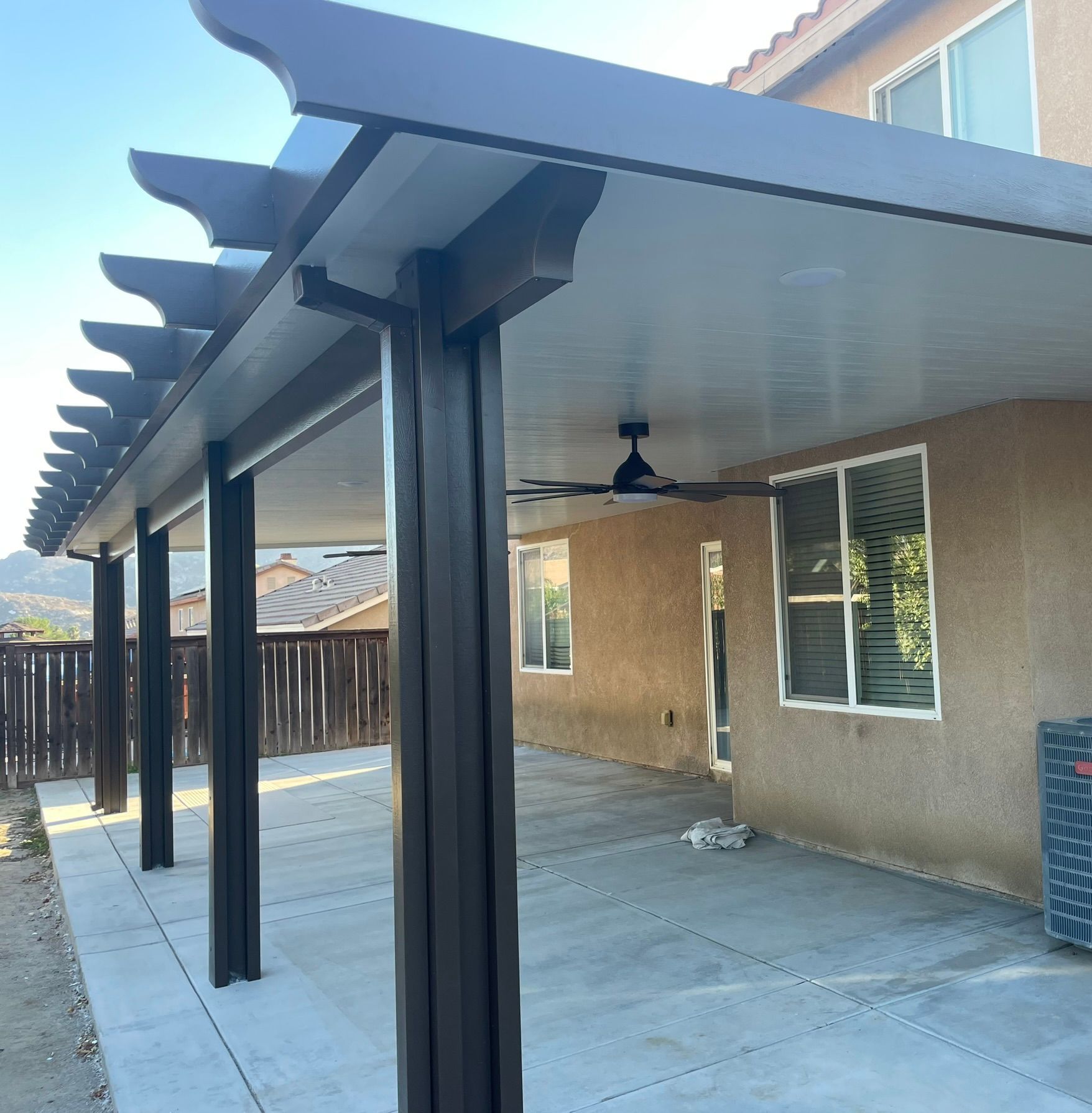 Covered patio with a dark brown pergola and ceiling fan, beside a beige house.