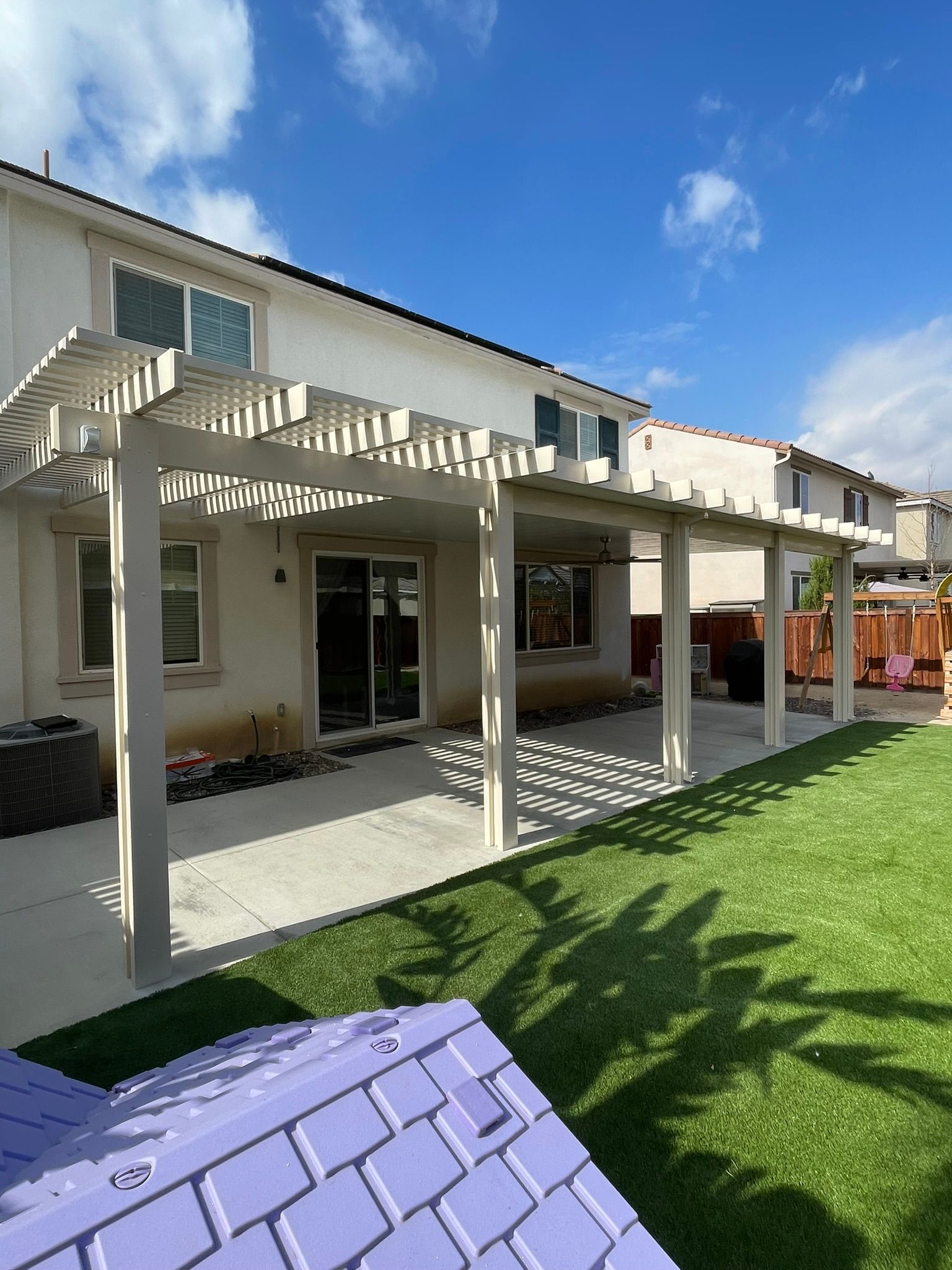 Backyard with a concrete patio, pergola, and green lawn; house with sliding glass door and blue sky.