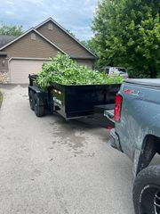 Gray pickup towing a black trailer piled high with green yard debris in a driveway