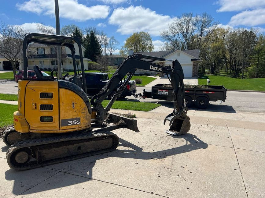 Yellow mini excavator on a concrete driveway beside a trailer, under a blue sky.
