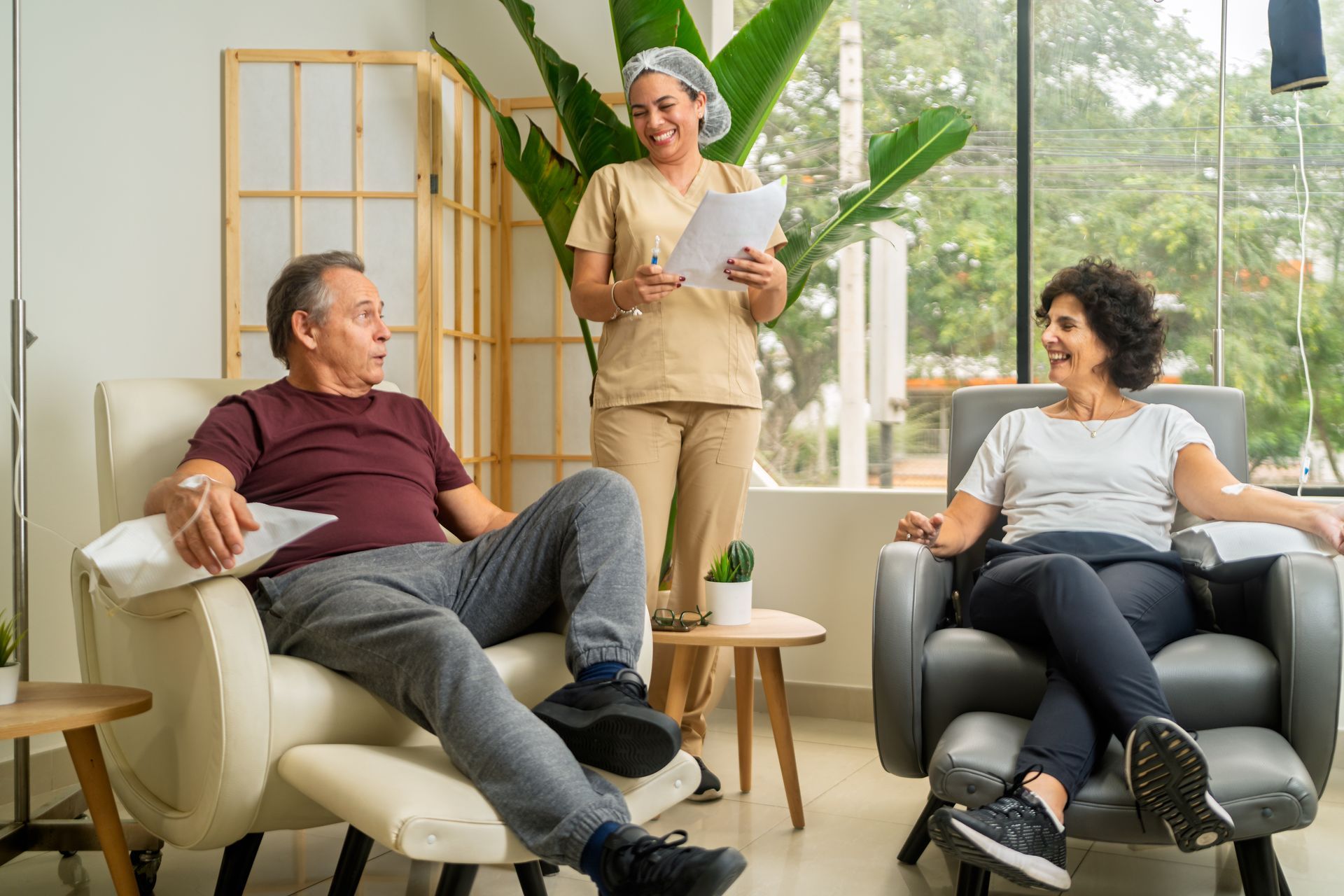 Two adults relax in chairs during wellness IV therapy while a smiling nurse reviews notes Two adults relax in chairs during wellness IV therapy while a smiling nurse reviews notes