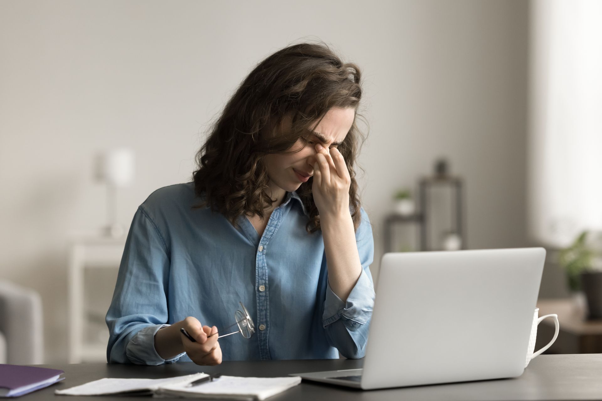 A person sitting at a desk with a laptop, holding glasses and rubbing the bridge of their nose in apparent eye strain.