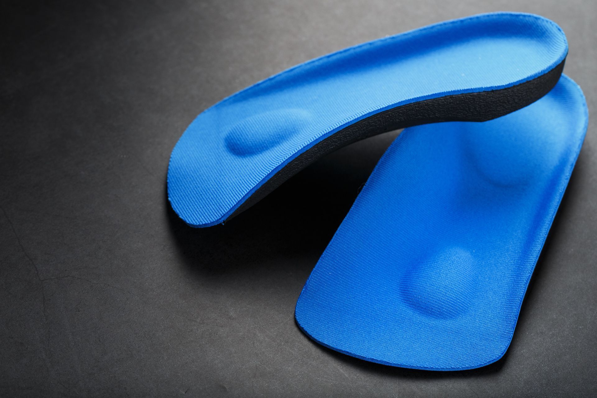 Two blue, contoured shoe insoles with raised arch support pads resting on a dark surface.