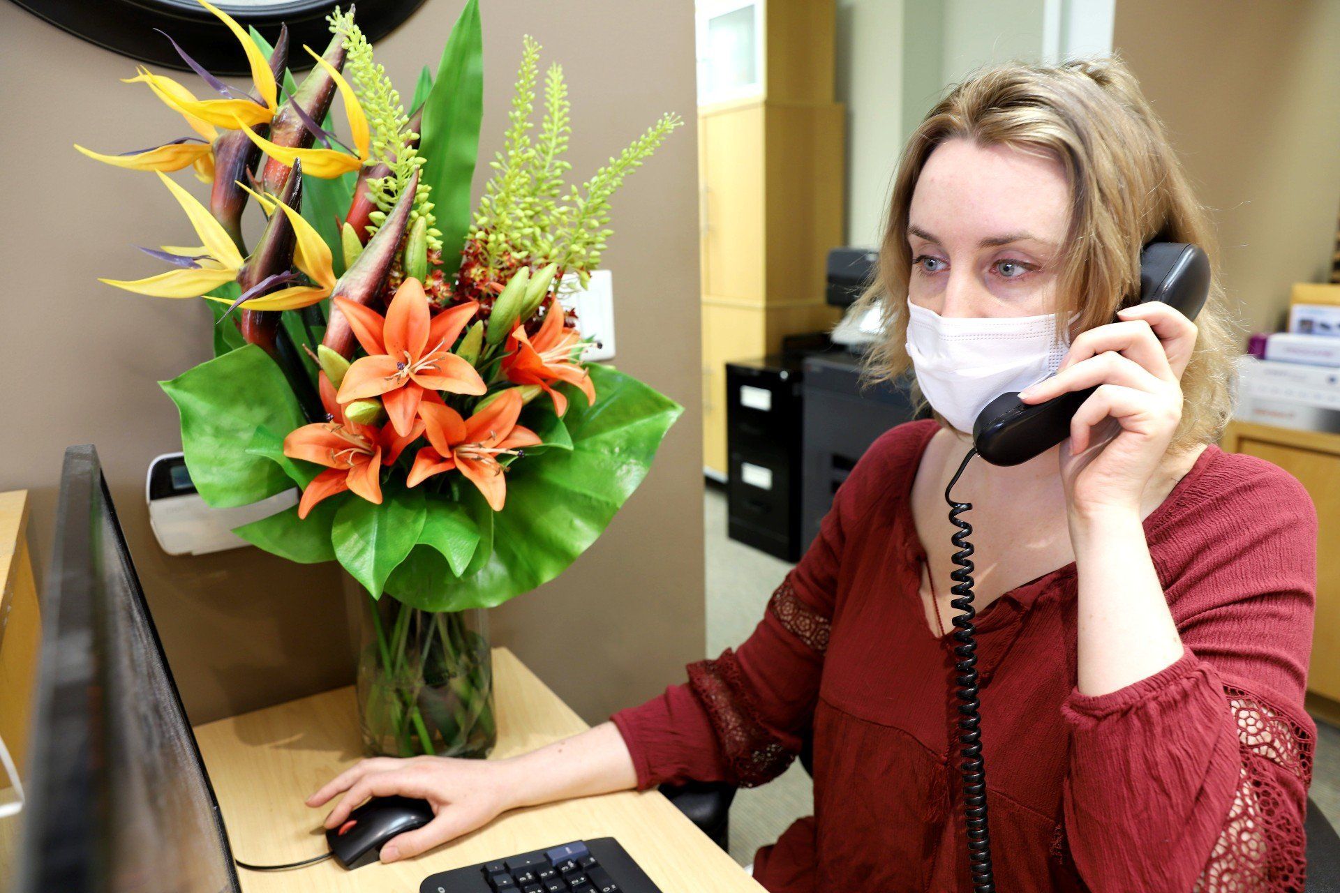 A woman wearing a mask is sitting at a desk talking on a phone.